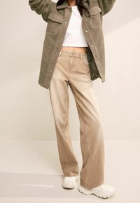Beige high-waisted jeans hebben een wijde pijp, met een zachte, gestructureerde afwerking. Gecombineerd met een witte crop top en een olijfgroene fleecejas.
