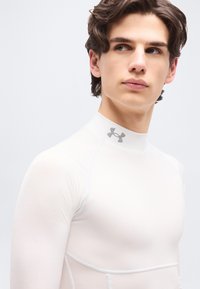 Νέος άνδρας με καστανά μαλλιά που φοράει άσπρο συμπιεστικό πουλόβερ με ψηλό λαιμό και γκρι λογότυπο Under Armour, κοιτάζοντας προς τα πλάγια.