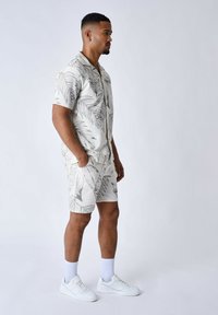 Camisa blanca de manga corta con un patrón de hojas grises, combinada con pantalones cortos a juego. Presenta un ajuste relajado y se lleva con zapatillas blancas y calcetines.