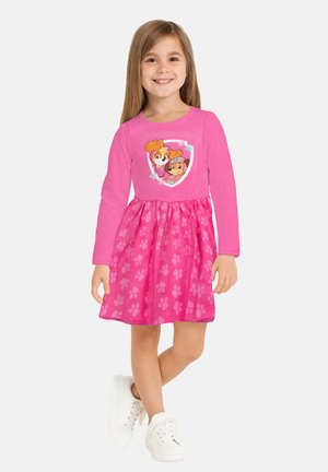 Bambina in un vivace vestito rosa con motivo di impronte di zampe e cani dei cartoni animati sul petto, che indossa sneaker bianche, in piedi su uno sfondo bianco.