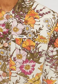 Giacca Wrangler con motivo floreale in crema, caratterizzata da fiori gialli, rosa e viola. Include bottoni di colore rame e due tasche sul petto.