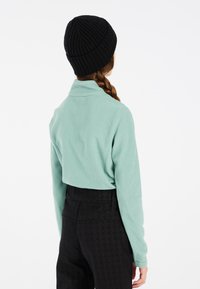 Maglione in pile verde menta chiaro con colletto ripiegato, abbinato a pantaloni neri a vita alta con motivo e un berretto nero a costine.