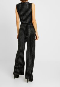 Femme vue de dos portant une combinaison sans manches noire avec des rayures verticales argentées et des chaussures à talons hauts noires se tenant sur un fond blanc.