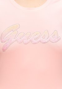 Κείμενο "Guesss" σε στυλ γραφής σε ανοιχτό ροζ ύφασμα διακοσμημένο με μικρά γυαλιστερά καρφάκια που σχηματίζουν τα γράμματα.