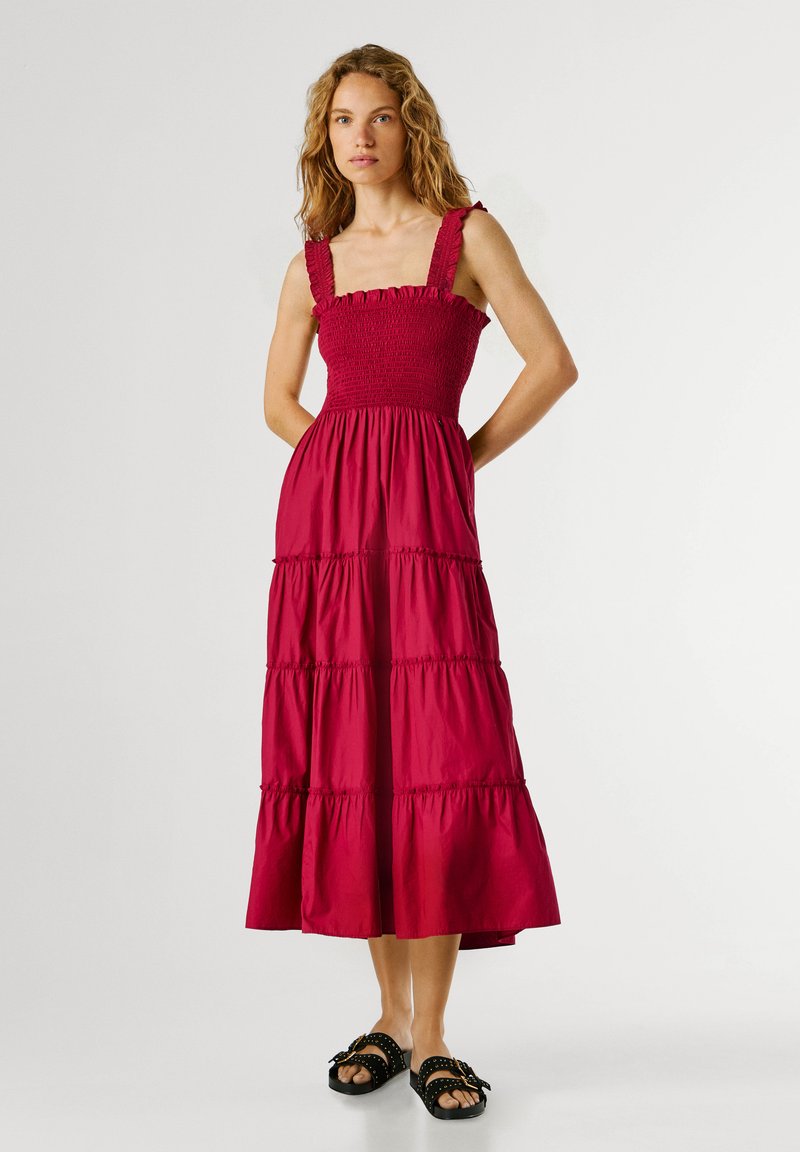 Donna con capelli mossi che indossa un vestito midi rosso senza maniche a balze e sandali neri slip-on borchiati, in piedi davanti a uno sfondo semplice.