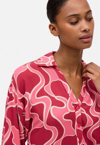 Blusa roja con un patrón ondulado en rosa y blanco roto, diseño con cuello, escote en forma de V y detalle de botones en la parte frontal. Textura de tela suave.