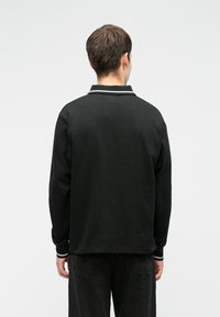 Persona vista da dietro che indossa una camicia nera a maniche lunghe con bordi bianchi su collo e polsini, e pantaloni scuri su uno sfondo semplice.