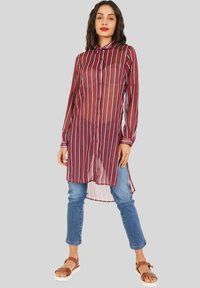 Camisa larga a rayas rojas y blancas, transparente, con cuello, botones y mangas largas. Combinada con jeans ajustados azules y sandalias marrones.
