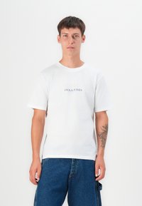 White cotton t-shirt with short sleeves, featuring a printed logo "JACK & JONES" in blue at the chest, paired with blue denim trousers.