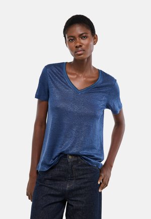 Femme portant un t-shirt bleu à col en V et manches courtes légèrement rentré dans un jean bleu foncé, posant avec une main sur la hanche.