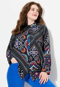Blusa nera a fantasia con disegni floreali e geometrici colorati. Maniche lunghe, colletto, con bottoni sul davanti. Abbinata a pantaloni blu.