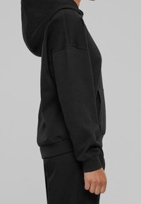 Persona con una sudadera negra con un bolsillo frontal y puños acanalados, vista de lado contra un fondo gris.