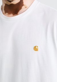 Persoon die een wit T-shirt draagt met een klein geel geborduurd logo op de borst, waarbij een deel van de nek en baard zichtbaar is.