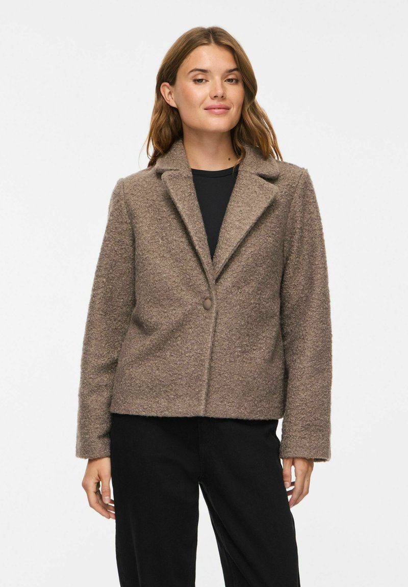 Femme portant une veste en laine marron texturée boutonnée sur un haut noir et un pantalon noir sur un fond blanc uni.