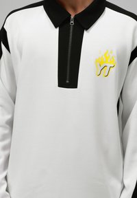 Polo cu mânecă lungă în alb și negru, prezentând un guler cu fermoar și un logo cu flacără galbenă cu "YT" pe piept. Textură moale, design sport.