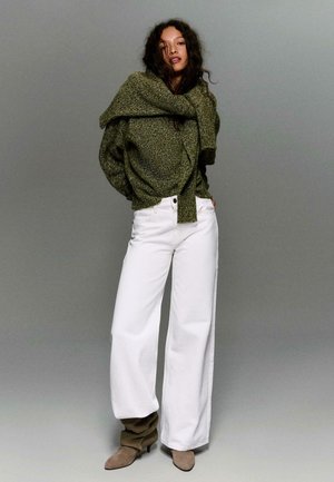 Žena s kudrnatými vlasy v olivově zeleném pleteném svetru a bílých širokých kalhotách stojící před jednobarevným šedým pozadím.