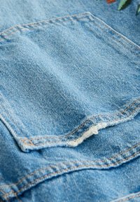 Svijetloplavi denimski materijal s ušivenom patch džepom. Vidljivo šivenje u smeđoj boji s istrošenim rubovima, teksturirano tkanje i lagano izbljeđivanje throughout.