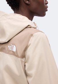 Vue de côté d'une personne portant une veste North Face beige et marron clair avec capuche, logo visible à l'arrière de l'épaule.