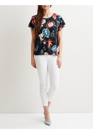 Musta kukkakuvioinen pusero, jossa on röyhelöhihat ja värikkäitä kasviaiheisia printtejä. Yhdistetty istuviin valkoisiin housuihin ja vaaleansinisiin korkoihin.