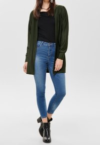 Langer olivgrüner Cardigan aus geripptem Stoff, kombiniert mit einem schwarzen Top und blauen Skinny-Jeans. Abgerundet mit schwarzen, schulterfreien Ankle-Boots.