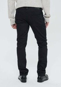 Personne portant un pantalon noir slim-fit, un pull gris clair moucheté et des chaussures de ville noires, se tenant de dos contre un fond uni.