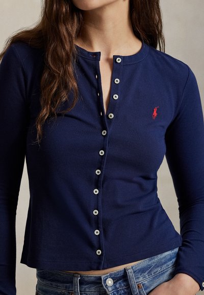 Sieviete tērpusies tumši zilā, garām piedurknēm ar pogām aprīkotā krekliņā ar sarkanu izšūtu polo spēlētāja logo un zilās džinsās.