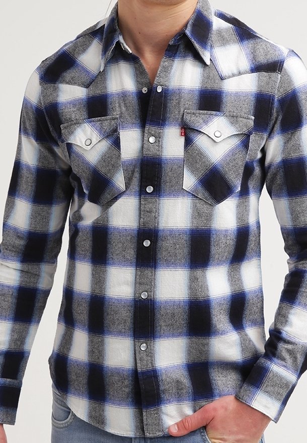 Homme portant une chemise à carreaux noire, blanche et bleue avec deux poches à rabat à l'avant, debout avec une main dans la poche du jean.
