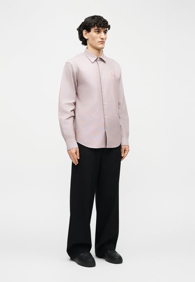 Νέος άνδρας με σγουρά μαλλιά, φοράει ανοιχτό ροζ πουκάμισο με κουμπιά και λογότυπο "EA", μαύρο παντελόνι με φαρδιά μπατζάκια και μαύρα παπούτσια, στέκεται μπροστά σε λευκό φόντο.