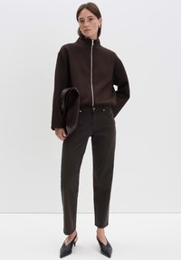 Vrouw in een donkerbruin ritsjack, zwarte broek, zwarte puntige pumps en met een zwart leren clutches tegen een effen achtergrond.