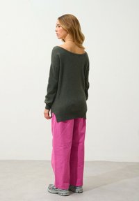 Übergroßer, dunkelgrüner Pullover im Off-Shoulder-Design, kombiniert mit leuchtend pinken, weit geschnittenen Hosen. Modell trägt graue Sportschuhe.