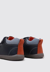 Zapatos de cuero azul marino con lengüeta de tela gris y tacón de ante rojo. Presenta una suela gris texturizada con pequeñas perforaciones. Diseñados para la comodidad.