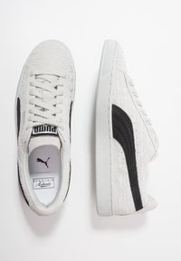 Valkoiset teksturoitu lenkkarit mustilla yksityiskohdilla, pyöreä kärki, litteä kumipohja ja merkittävä logo kielellä; suunnittelussa korostuu minimalistisuus ja mukavuus.