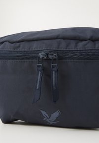 Sötétkék nylon utazótáska, texturált felülettel, dupla cipzárral és egy oldalán dombornyomott sas logóval.