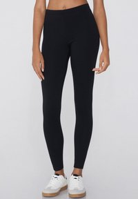 Leggings noirs en tissu extensible avec une taille haute, présentant une texture lisse et un design ajusté. Portés avec des baskets blanches.