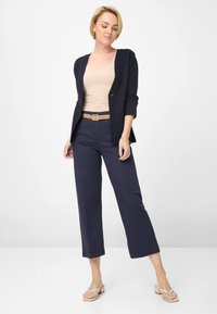 Marineblazer mit Dreiviertelärmeln, beigem Tanktop, marineblauen weitem Hosen und einem hellbraunen Gürtel mit einer Metallnadel. Helle Sandalen.