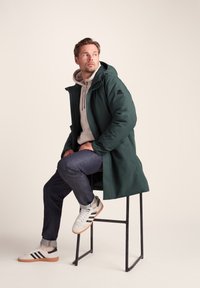 Green waterproof coat with a hood, worn over a light gray hoodie, paired with dark jeans and white sneakers featuring black stripes.