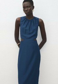 Žena s krátkymi vlasmi má na sebe bezrukávové modré šaty so bočnými výstrihmi, zlaté kruhové náušnice a náramok, stojí pred rovnou bielou stenou.