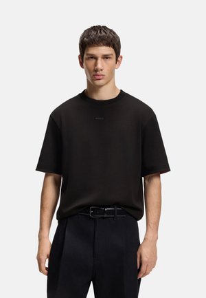 Young man with short brown hair wearing a plain black short-sleeve T-shirt tucked into black pants with a black leather belt.