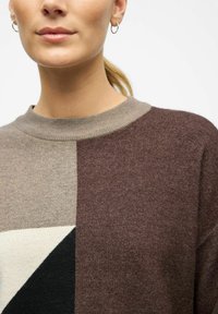 Brun og beige striktrøje med geometrisk design, der har en rund halsudskæring og forskellige teksturer, inklusive ensfarvede og mønstrede sektioner.