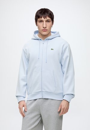 Junger Mann trägt hellblauen Kapuzenpullover mit Reißverschluss und graue Jogginghose, steht vor weißem Hintergrund.