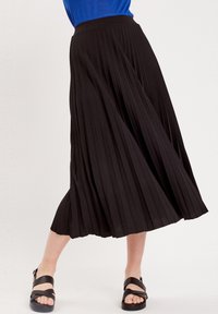 Jupe plissée noire à taille haute. Tissu léger avec une texture douce. Associée à des sandales noires. La jupe arrive au milieu du mollet.
