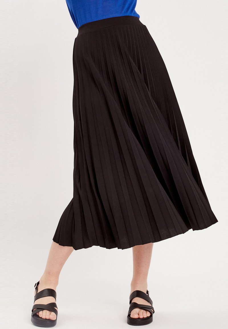 Jupe plissée noire à taille haute. Tissu léger avec une texture douce. Associée à des sandales noires. La jupe arrive au milieu du mollet.