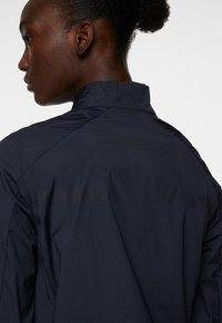 Marinblå vindjacka med hög krage och texturerad baksida. Har ett slätt tyg med ett diskret logomönster som visar sömmar och detaljer.