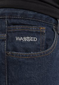 Sötét farmernadrág sárga varrással, hátsó zsebbel és fehér cérnával hímzett "WASTED" logóval. Simaság jellemzi.