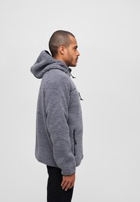 Sudadera de forro polar gris con capucha, bolsillos laterales con cremallera y material texturizado. Presenta un ajuste holgado y cordones ajustables.