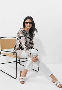 Langärmliges Oberteil mit abstraktem Design in Schwarz und Pink, kombiniert mit weißen Hosen und weißen Rutschen-Sandalen, sitzend auf einem schwarz-gerahmten Stuhl.