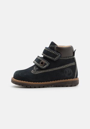 Botte noire pour tout-petit avec double fermeture velcro, coutures blanches, col rembourré à la cheville et semelle en caoutchouc marron sur un fond clair uni.