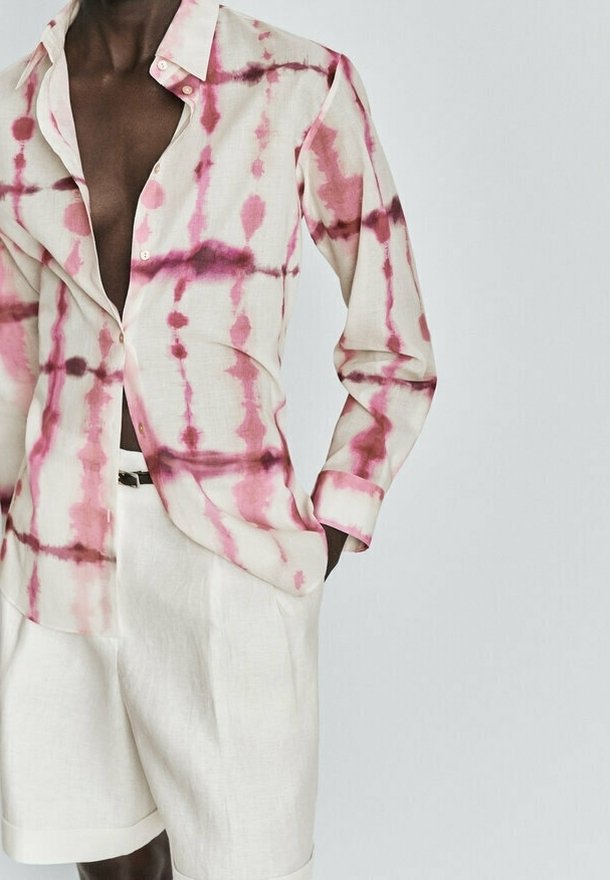 Homme portant une chemise tie-dye manches longues ouverte rose et blanche avec un short blanc, debout avec une main dans la poche sur un fond uni.