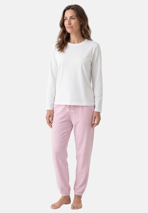 Kobieta stojąca boso, ubrana w białą koszulę z długim rękawem i jasnoróżowe spodnie dresowe z sznurkiem, na jednolitym tle.