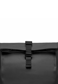 Sac à dos noir à rabat avec une finition texturée, doté d'une boucle centrale et d'une sangle réglable pour une fermeture sécurisée. Un design simple et fonctionnel.